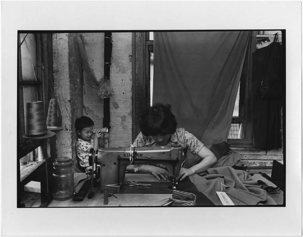 A woman sewing at a garment factory on Canal Street with a child sitting behind her. 1979-1982. Courtesy of Paul Calhoun, Museum of Chinese in America (MOCA) Collection